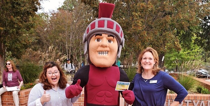 Three people pose cheerfully for a photo outdoors on a brick courtyard. In the center is a person in a Trojan mascot costume with a maroon helmet and outfit, giving two thumbs up.